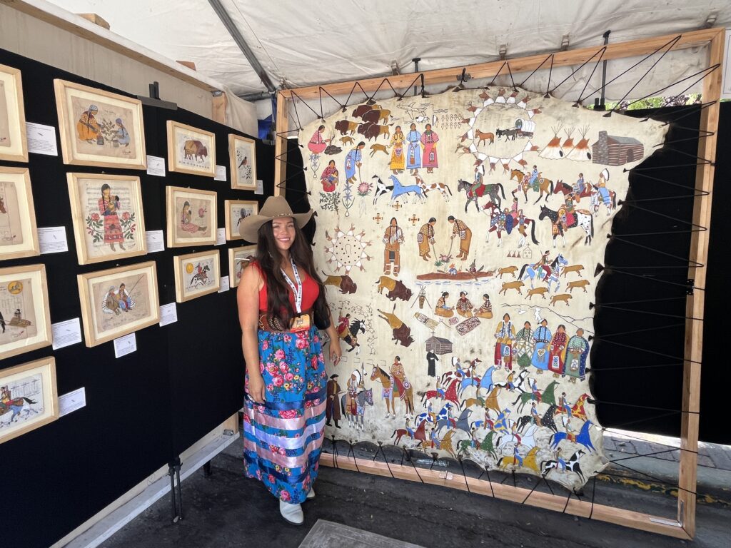 Aspen Decker (Confederated Salish and Kootenai Tribes–T̓ata̓yáqn, Ql̓isp̓é & Ksanka) standing alongside her pictorial painting on buffalo hide depicting the story of the Inland Salish people.
